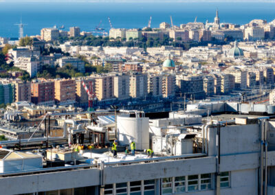 Operai sul tetto dell'Ospedale San Martino durante l'installazione ISAAC, con Genova e il porto sullo sfondo
