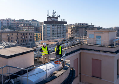 Due operai sul terrazzo del Padiglione Specialità accanto a un dispositivo ISAAC coperto, Genova sullo sfondo